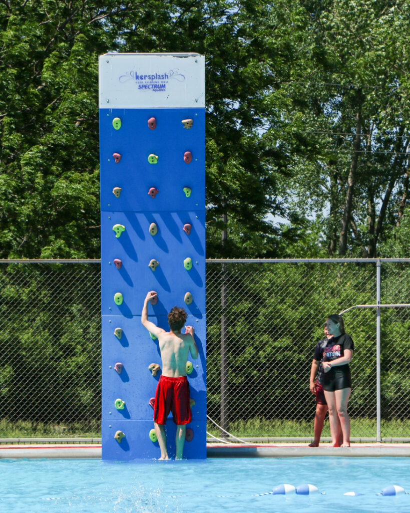 A boy wearing red swimming trunks climbs the pool climbing wall.